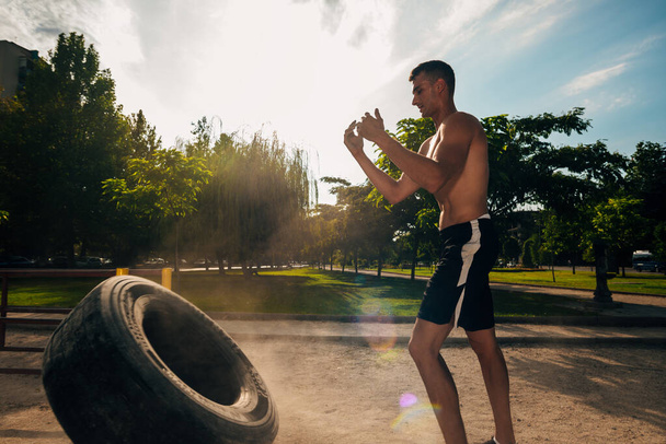 Muscular fitness shirtless man moving large heavy tire in street gym. Concept lifting, workout training - Photo, Image
