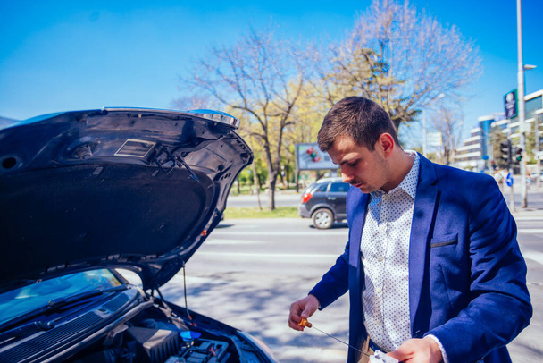 Un hombre de negocios ha aparcado su coche al lado del bulevar mientras revisa su aceite de coche, en un día soleado y ajetreado
. - Foto, Imagen