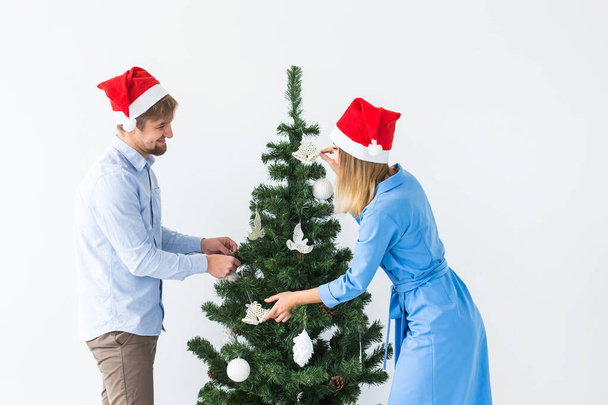 Holidays and festive concept - Young family couple in santa hats decorating the christmas tree - 写真・画像