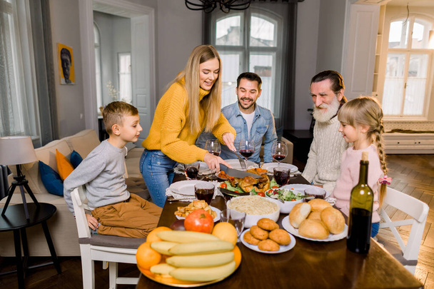 Family of five people, grandfather, parents and children sitting at the table and going to eat roasted turkey, while happy mother is cutting turkey. Thanksgiving family dinner concept - Photo, Image