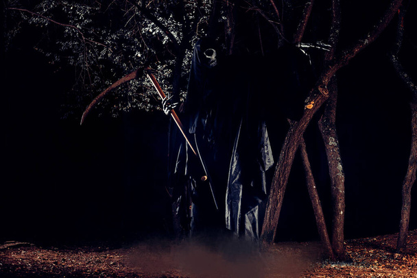 Retrato de sombrío segador, el ángel de la muerte (Azrael) acechando por el bosque por la noche rodeado de niebla, humo en un bosque místico sosteniendo su tiña. - Foto, imagen
