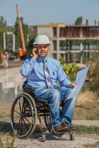 Vammainen henkilö rakennuskypärässä, jolla on kädessään asiakirjoja, jotka puhuvat puhelimessa rakennuksen taustalla. Onnistunut pyörätuolin käyttäjä ohjaa työtä
. - Valokuva, kuva