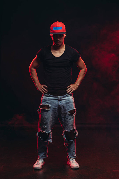 portrait of nice vigorous man wearing black t-shirt, jeans and cap isolated over red smoky background - Photo, Image
