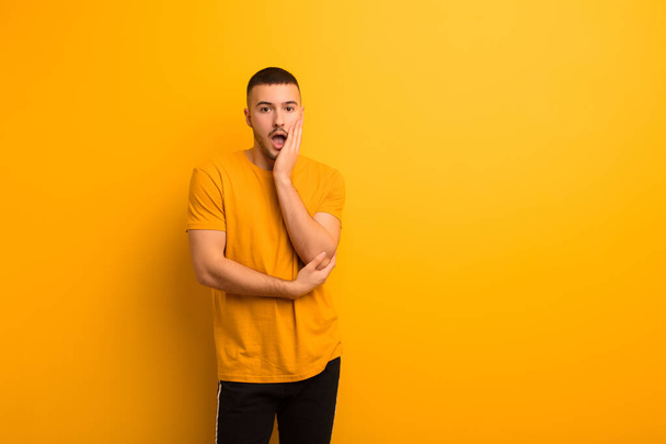 young handsome man open-mouthed in shock and disbelief, with hand on cheek and arm crossed, feeling stupefied and amazed against flat background - Fotoğraf, Görsel