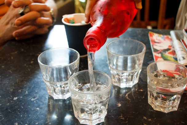 Nahaufnahme Hand gießt Wasser aus einer roten Flasche in Glasgläser auf einem Restauranttisch - Foto, Bild