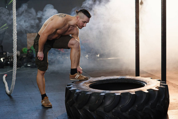 Muscular man with naked torso has a rest near heavy tire on the smoke and sunlight background in the gym. - Fotografie, Obrázek
