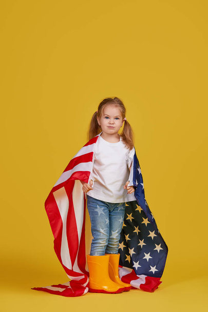 Patriotic holiday. Happy child, cute little girl with american flag on yellow studio background. USA celebrate July 4th, independence Day. - 写真・画像