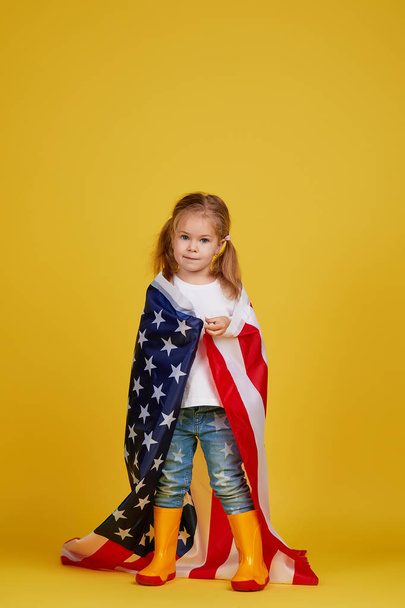 Πατριωτική γιορτή. Χαρούμενο παιδί, χαριτωμένο κοριτσάκι με αμερικάνικη σημαία σε κίτρινο φόντο στούντιο. Η Ούσα γιορτάζει την 4η Ιουλίου, Ημέρα ανεξαρτησίας. - Φωτογραφία, εικόνα