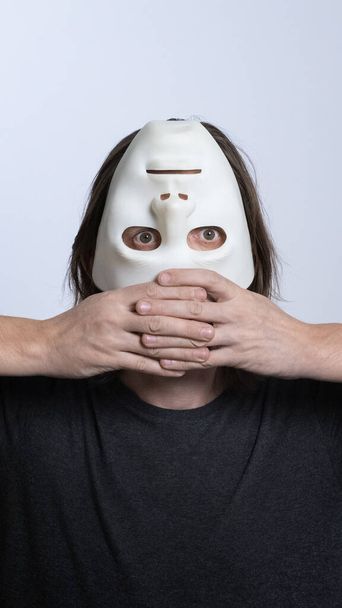 Ein langhaariger Mann hält eine weiße Maske kopfüber in der Nähe seines Gesichts. das Konzept des Versteckens einer Person, des Guckens, psychischer Probleme, einer gespaltenen Persönlichkeit, Psychedelie oder Voodoo-Rituals. vertikaler Rahmen. - Foto, Bild