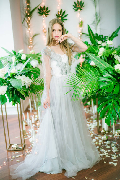 Attractive young girl with blond hair and evening make-up in a romantic dress among tropical greenery and white orchids, roses and lilies. Beauty and fashion - Photo, Image