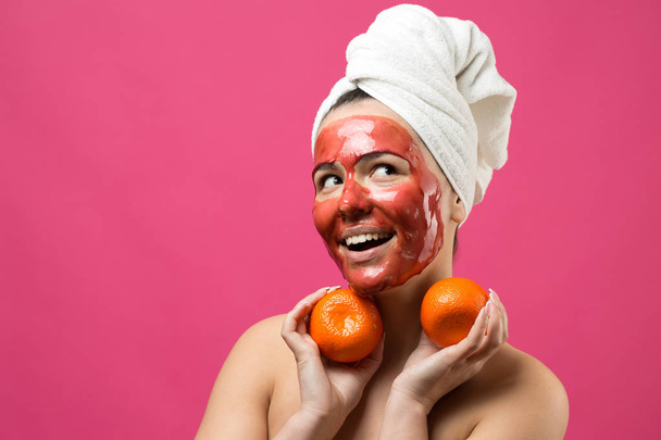 Retrato de beleza de mulher em toalha branca na cabeça com máscara nutritiva vermelha no rosto. Skincare limpeza eco orgânico spa cosmético relaxar conceito. Uma menina fica de costas segurando um mandarim laranja
. - Foto, Imagem