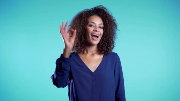 Positive young african girl smiles to camera. Young woman making OK sign over blue background. Winner. Success. 4k - Séquence, vidéo