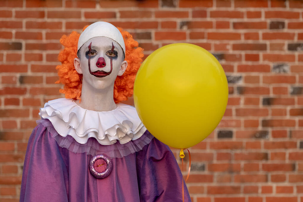 Un tizio in costume da clown con un palloncino giallo contro un muro di mattoni. Costume per tutti i santi giorno carnevale o Halloween
. - Foto, immagini