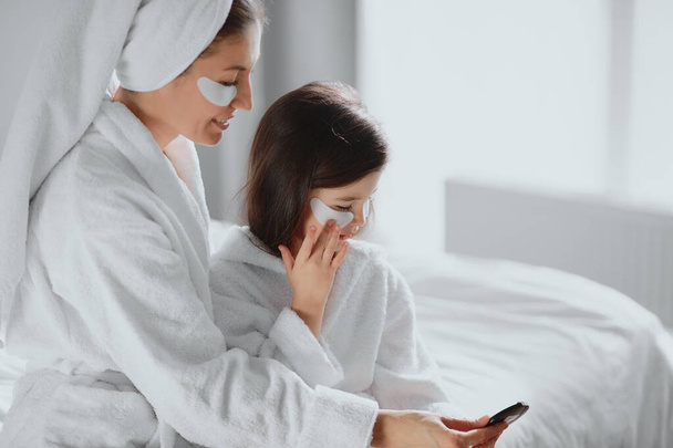 awesome mom and daughter sit together on bed in beauty masks patches at home - Fotoğraf, Görsel