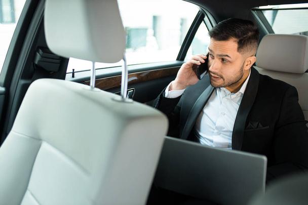 Nuori liikemies työskentelee kannettavassa tietokoneessa ja puhuu älypuhelimella autossa
 - Valokuva, kuva