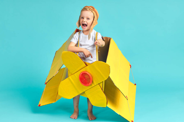 Kid boy playing with yellow cardboard airplane isolated on blue - Фото, изображение