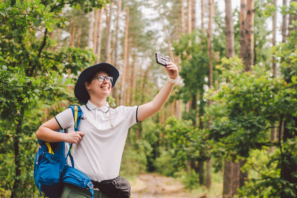 Sorrindo turista feminina detém telefone inteligente, leva selfie contra bela madeira paisagem. Mulher caminhando com mochila tirando foto selfie com smartphone. Viagens e estilo de vida saudável ao ar livre
 - Foto, Imagem