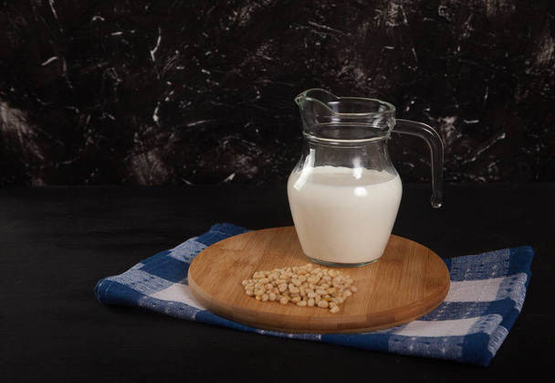 Pine nut vegetable milk in a glass jug stands on a wooden stand on a black background - Foto, afbeelding