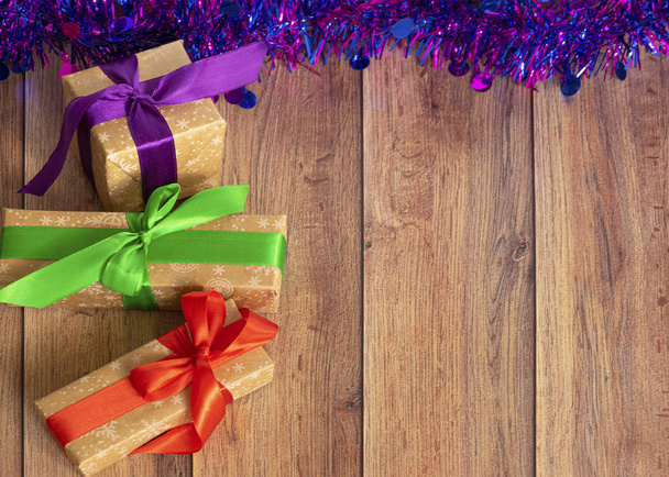 Τα δώρα είναι συσκευασμένα σε χαρτί Kraft και δεμένα με σατέν κορδέλα με μωβ tinsel σε ξύλινο φόντο. - Φωτογραφία, εικόνα