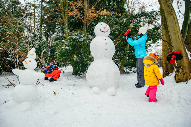 Kar parkında kardan adam yapan genç bir aile. Küçük kız yardım ediyor. - Fotoğraf, Görsel