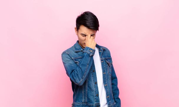 young chinese man feeling stressed, unhappy and frustrated, touching forehead and suffering migraine of severe headache against flat color wall - Foto, imagen