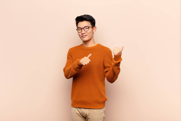 young chinese man smiling cheerfully and casually pointing to copy space on the side, feeling happy and satisfied against flat color wall - Фото, изображение