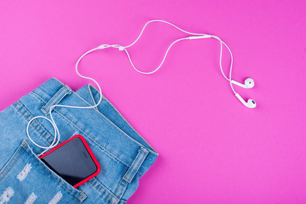 青いジーンズ、バックポケットにピンクの背景の音楽のためのヘッドフォン付きの電話. - 写真・画像