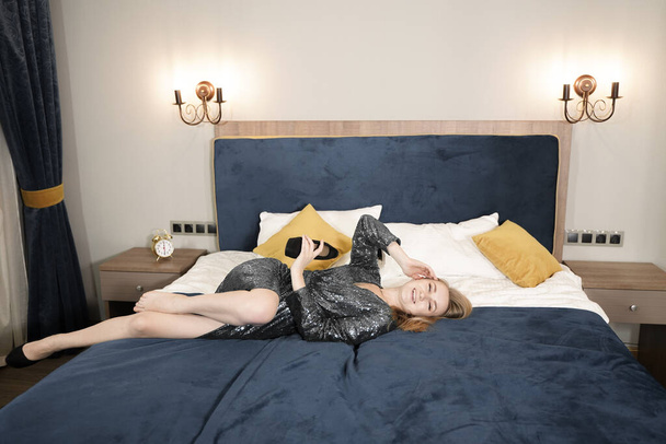 junge Frau im eleganten schwarzen Abendkleid im Bett liegend. Erwachsene kaukasische blonde Mädchen in Pailletten Outfit entspannen im Schlafzimmer. - Foto, Bild