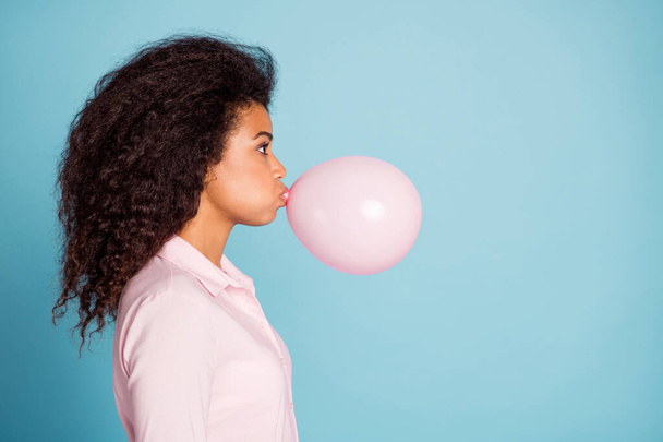 Closeup profile photo of pretty dark skin lady holding big air balloon in mouth making decorations for surprise party wear pink shirt isolated blue color background - Photo, Image