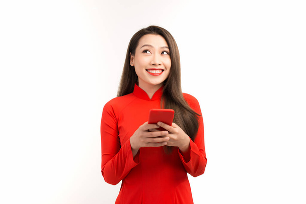 Portrait of beautiful Asian woman using mobile phone and wearing red Ao dai on white background. - Valokuva, kuva