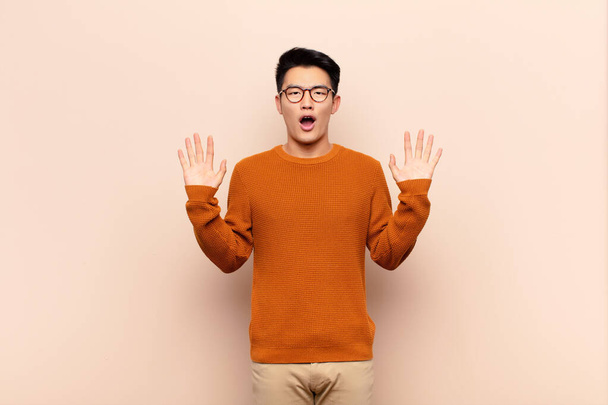 young chinese man feeling stupefied and scared, fearing something frightening, with hands open up front saying stay away against flat color wall - Fotografie, Obrázek