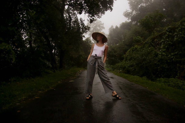 Mladá žena v tradičním bambusovém klobouku a širokých kalhotách na cestách, období dešťů na tropickém ostrově, turistika ve vzdálených destinacích - Fotografie, Obrázek