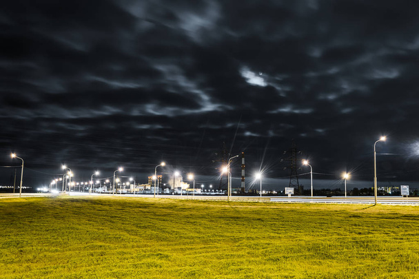 La route de nuit est éclairée par des lumières vives et des vitesses d'obturation. Usine avec tuyaux d'échappement. Lune et étoiles, nuages flous et herbe verte luxuriante. Cartouche : "Pour des routes propres, Nachalovo, Krasny Yar, Atyrau
." - Photo, image