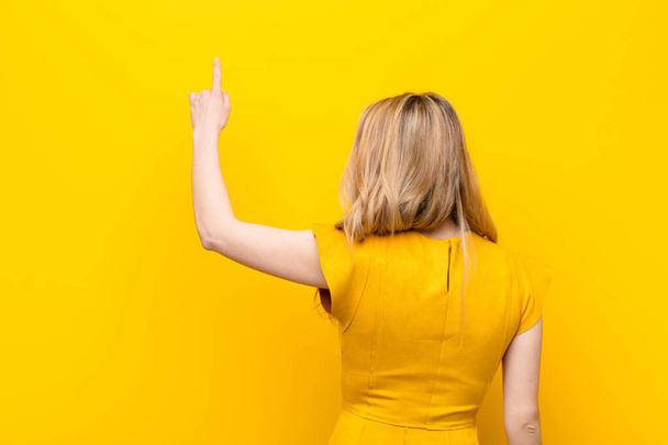 young pretty blonde woman standing and pointing to object on copy space, rear view against flat color wall - Фото, изображение