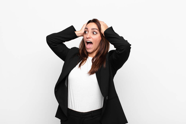 young pretty businesswoman with open mouth, looking horrified and shocked because of a terrible mistake, raising hands to head against white wall - Fotoğraf, Görsel
