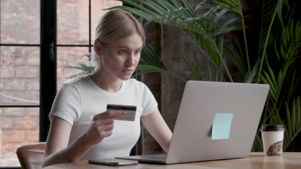 Pretty girl is making online payment holding bank card using modern laptop at home sitting and smiling. Finance, shopping in internet and youth concept. - Materiał filmowy, wideo