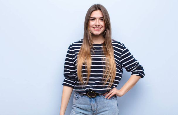 young pretty woman smiling happily with a hand on hip and confident, positive, proud and friendly attitude against soft blue wall - Fotoğraf, Görsel