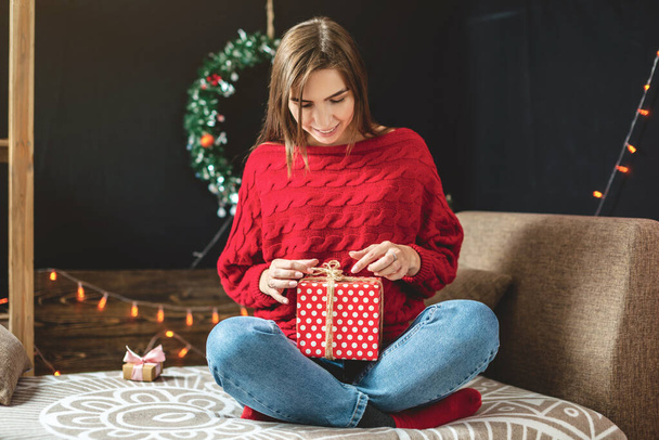 Woman in red sweater sits on a sofa with a gift box in her hands on background of garland lights. Cozy Christmas mood - Zdjęcie, obraz