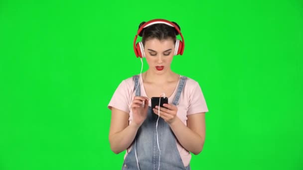 Girl in big red headphones chooses music on mobile phone on green screen - Filmati, video