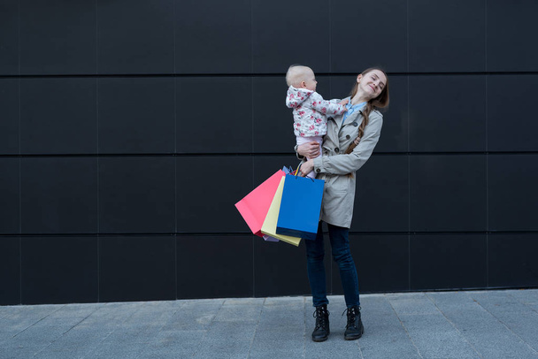 Young mother with little daughter on the arms and shopping bags in hand. Black street wall on background - Photo, Image