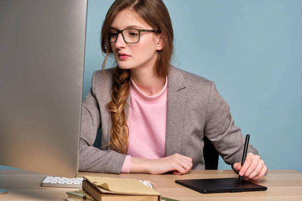 A girl with a braid in a gray jacket and glasses sits at a table and looks at the monitor, working on a graphics tablet. - 写真・画像