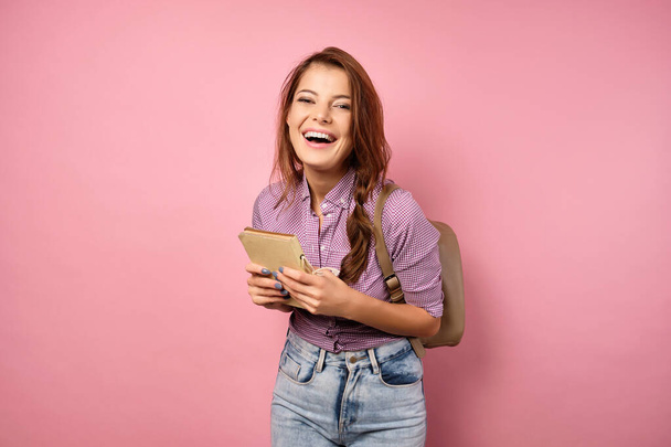 A brunette with a braid stands on a pink background in a plaid shirt and jeans with a book and a backpack, laughing - Φωτογραφία, εικόνα