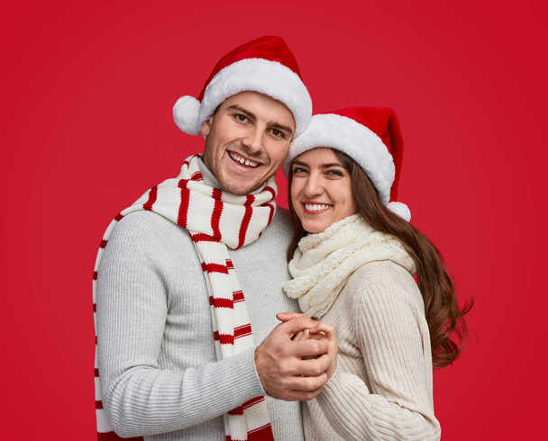 Wesoła para w czerwonym świątecznym kapeluszu i szaliku przytula się i uśmiecha do kamery - Zdjęcie, obraz