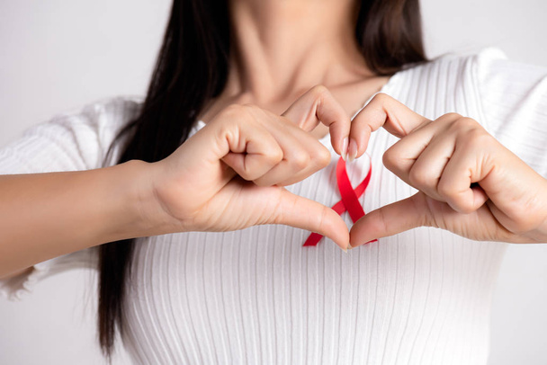 Closeup of woman hand in shape of heart with red badge ribbon on chest to support AIDS Day. Healthcare, medicine and AIDS awareness concept. - Foto, Bild