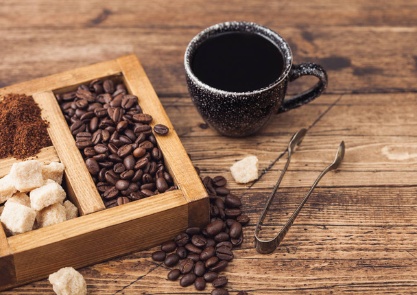 Black ceramic cup of fresh raw organic coffee with beans and ground powder with cane sugar in vintage box on wooden background. Top view - Foto, Imagen