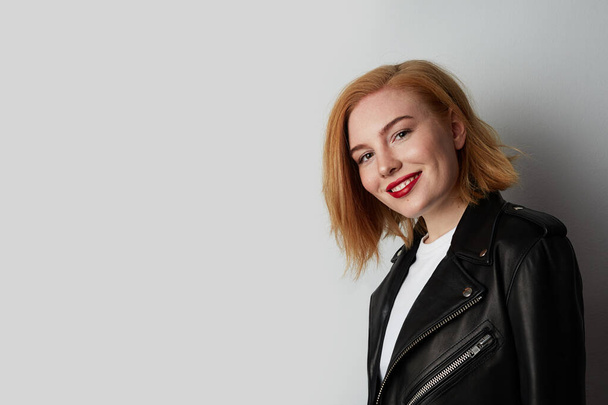 Portrait of young woman with red hair posing in the studio and wearing leather biker jacket. Space for text. - Fotó, kép