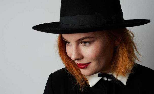 Portret hipsterskiej młodej kobiety w czarnej sukience i kapeluszu pozującej na białej ścianie. Izolacja. - Zdjęcie, obraz