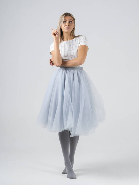 Vertical shot of amazed blonde girl model have an idea on cyclorama white background in a studio. Hand with finger pointing up. Full growth in profile in pastel colors. Long skirt and gray tights - Foto, Imagen
