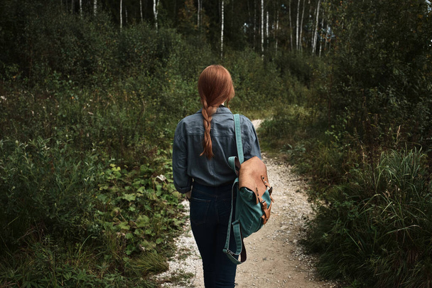 A girl with a red braid in a denim shirt with a backpack walks along a path in the middle of the forest, a frame from the back. - 写真・画像