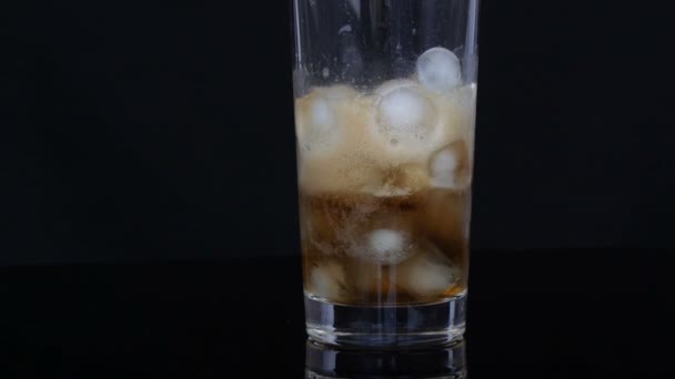 Cola is poured into a large long glass cup with pieces of ice inside on a black background. - Footage, Video